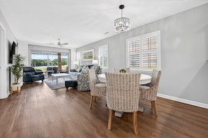 A spacious living room with a large window, a dining area with wicker chairs, and a ceiling fan. - 21d Beaches & Birdies 3 Bedroom Home (Pawleys Island)