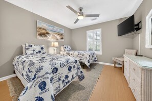 Quarto com duas camas, ventilador de teto, televisão e uma pintura com tema praiano.