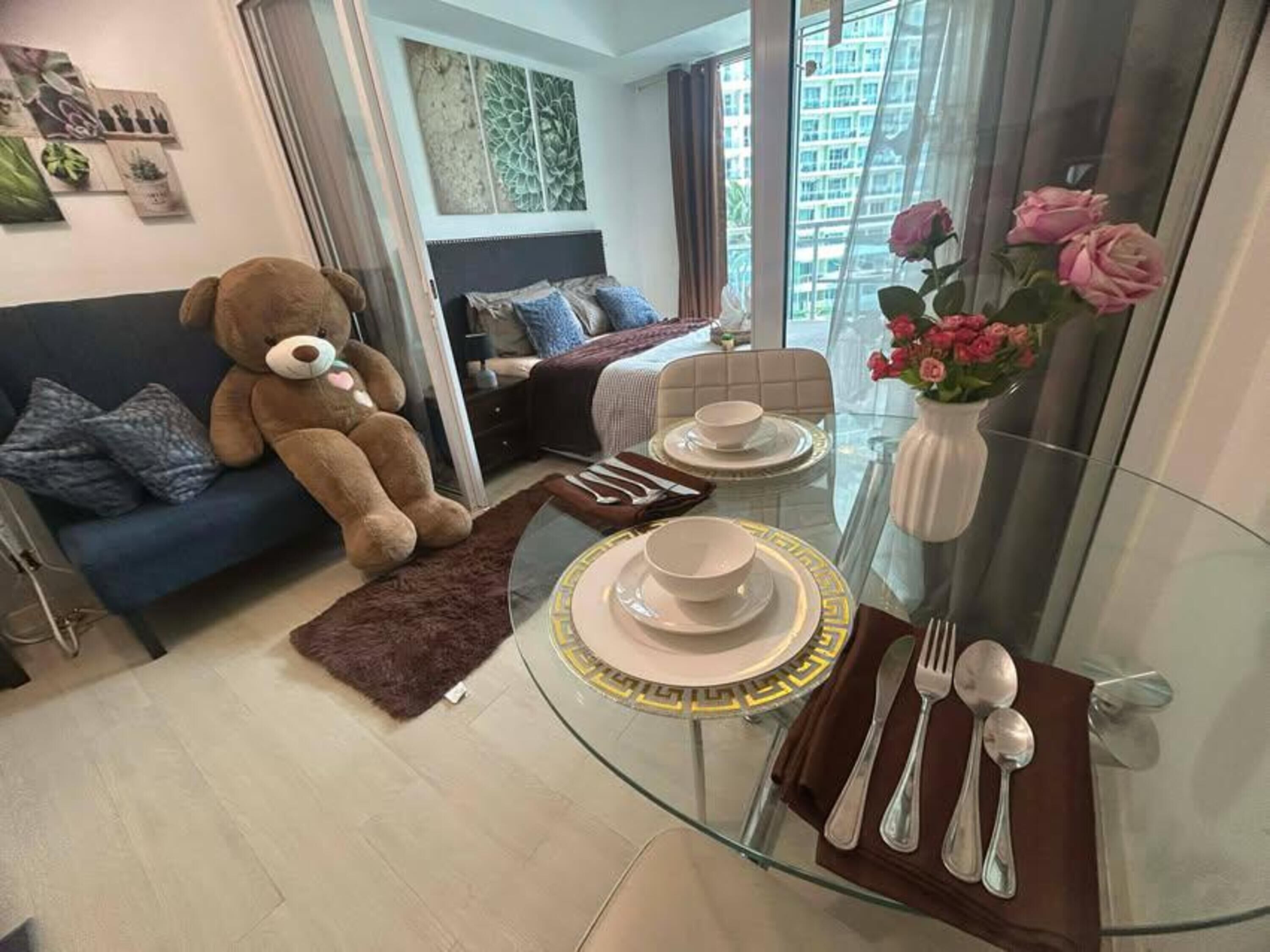 A modern living room with a glass dining table set for two, a sofa with a teddy bear, and a vase of flowers.