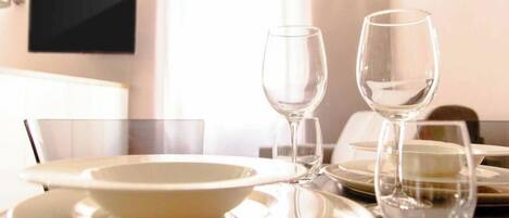 A dining table set with white plates, bowls, and wine glasses, with a television mounted on the wall in the background.