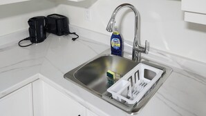 A kitchen with white cabinets, a stainless steel sink, a wooden shelf with a plant and a decorative sign, and a bottle of Dawn dish soap. - Beautiful Oasis Haven 1 Bedroom unit (Moncton)