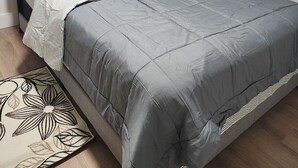 A neatly made bed with grey bedding, a bedside table with a lamp, and a small potted plant on the windowsill. - Beautiful Oasis Haven 1 Bedroom unit (Moncton)