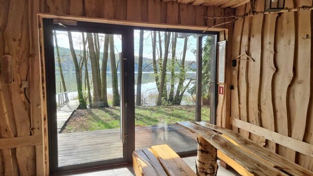 Una cabaña de madera con un amplio ventanal de vidrio que da a una terraza con vistas a un bosque y un cuerpo de agua.