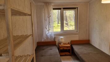 A small, single-room cabin with a window providing a view of a forest. There is a wooden bed with a headboard, a small table with a lamp, and a radiator under the window.