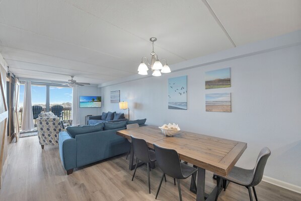A modern living room with a wooden dining table, blue sofa, and a view of the ocean through large windows. - Enjoy the sun rise in the morning and breath in the sea air during the day on th (Port Aransas)