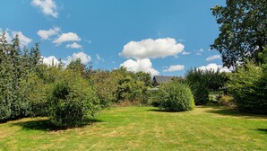 Un champ verdoyant, parsemé d’arbustes denses et d’arbres, un ciel bleu clair avec quelques nuages épars, et un bâtiment avec un toit en pente à l’arrière-plan.