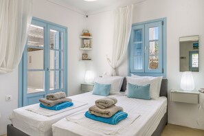 A hotel room with two beds, blue-framed windows, white walls, and a bedside table with a lamp.