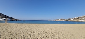 A sandy beach with a clear blue sea, white buildings on the hills, and a wooden structure on the left.