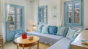 A room with a blue-framed window, a sofa with striped cushions, a round wooden table with a red basket, and a lamp. - Beachfront Ios Villa (Ios)