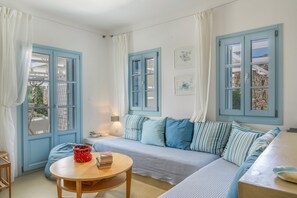 A room with a blue-framed window, a sofa with striped cushions, a round wooden table with a red basket, and a lamp.