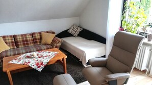 A compact living space with a sofa, a small table with a floral tablecloth, a chair, and a bed. - Apartment Ingrid Alfes, Apartment 1 (Lennestadt-Halberbracht)