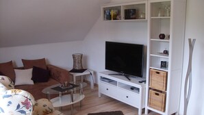 Un salon comprenant un canapé, une table basse, un téléviseur posé sur un meuble, une bibliothèque et un lampadaire.