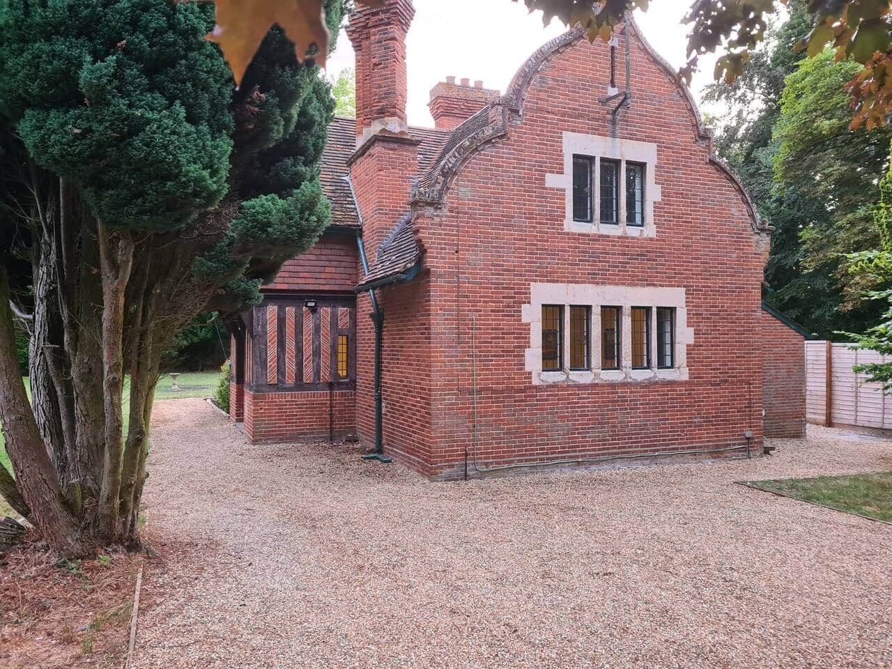 A brick house with a small attached structure, a gravel driveway, and a large tree in the foreground.