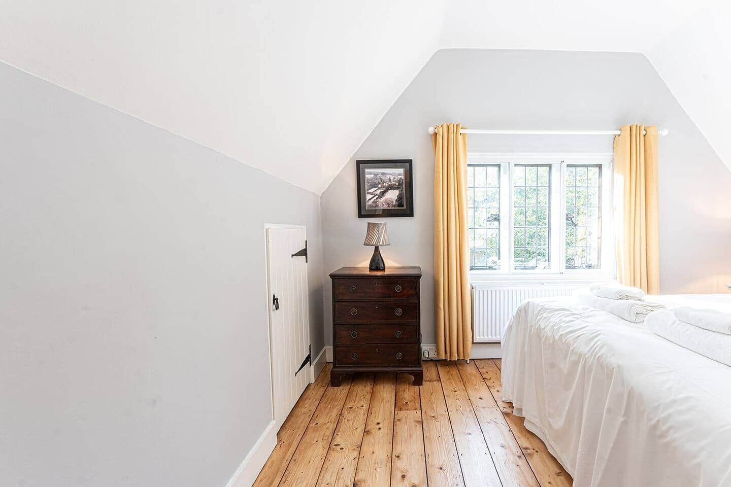 Una camera da letto con pavimento in legno, un letto bianco, una cassettiera, un quadro incorniciato e una finestra con tende.