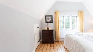 Una camera da letto con pavimento in legno, un letto bianco, una cassettiera, un quadro incorniciato e una finestra con tende.