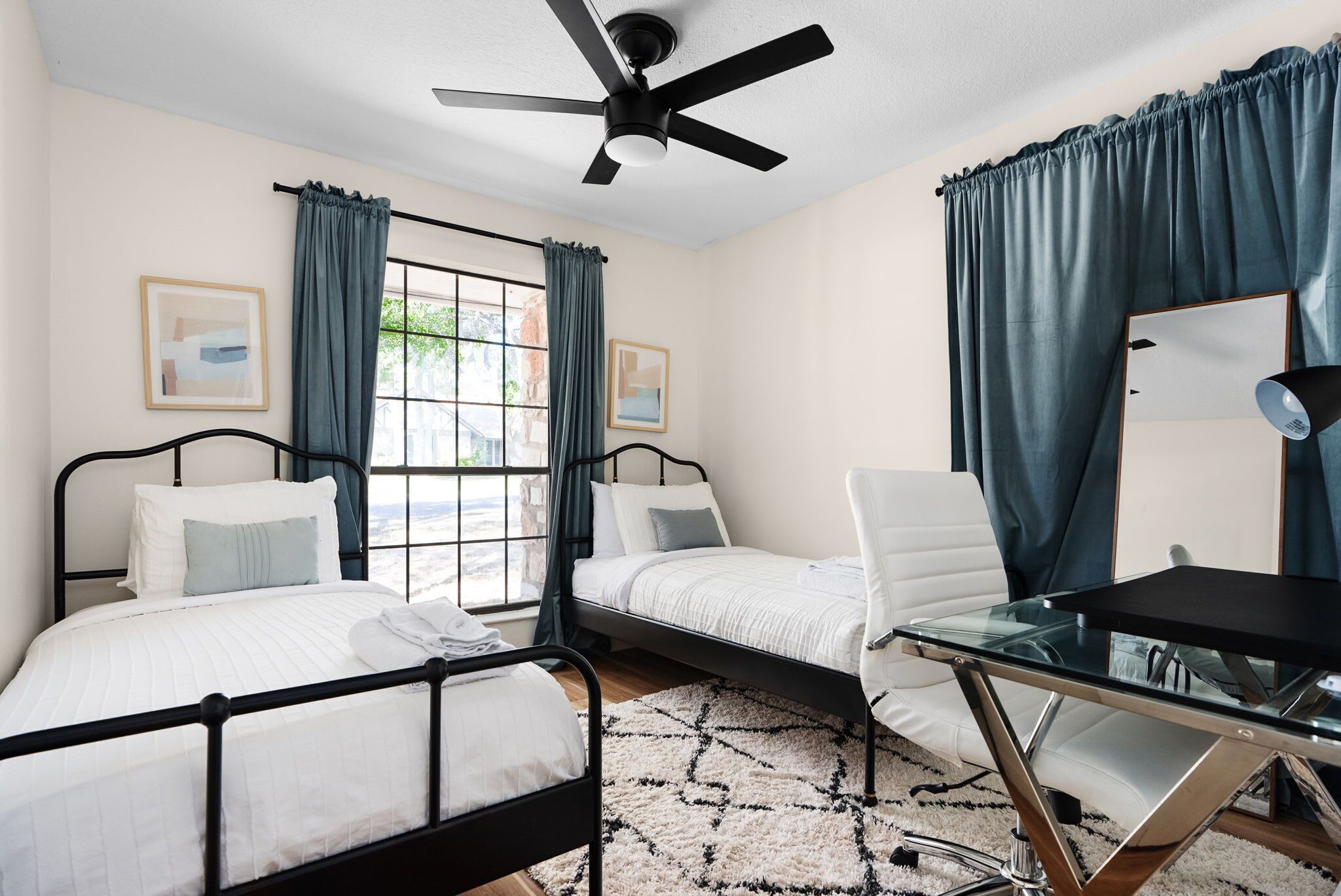 A bedroom with two beds, a desk, a chair, a ceiling fan, and a window with curtains.