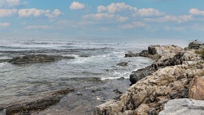 A rocky shoreline with waves, clear sky, and scattered clouds.