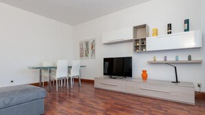 A modern living room with a grey sofa, a glass dining table with chairs, a flat-screen TV, a coffee table, and built-in shelves with various items. - Modern house in Mestre with direct streetcar to Venice (Venezia)