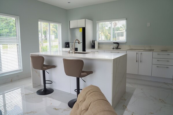 A modern kitchen with a marble countertop, two bar stools, a central island, and a built-in oven. - Paradise Suite (Cotton Ground)