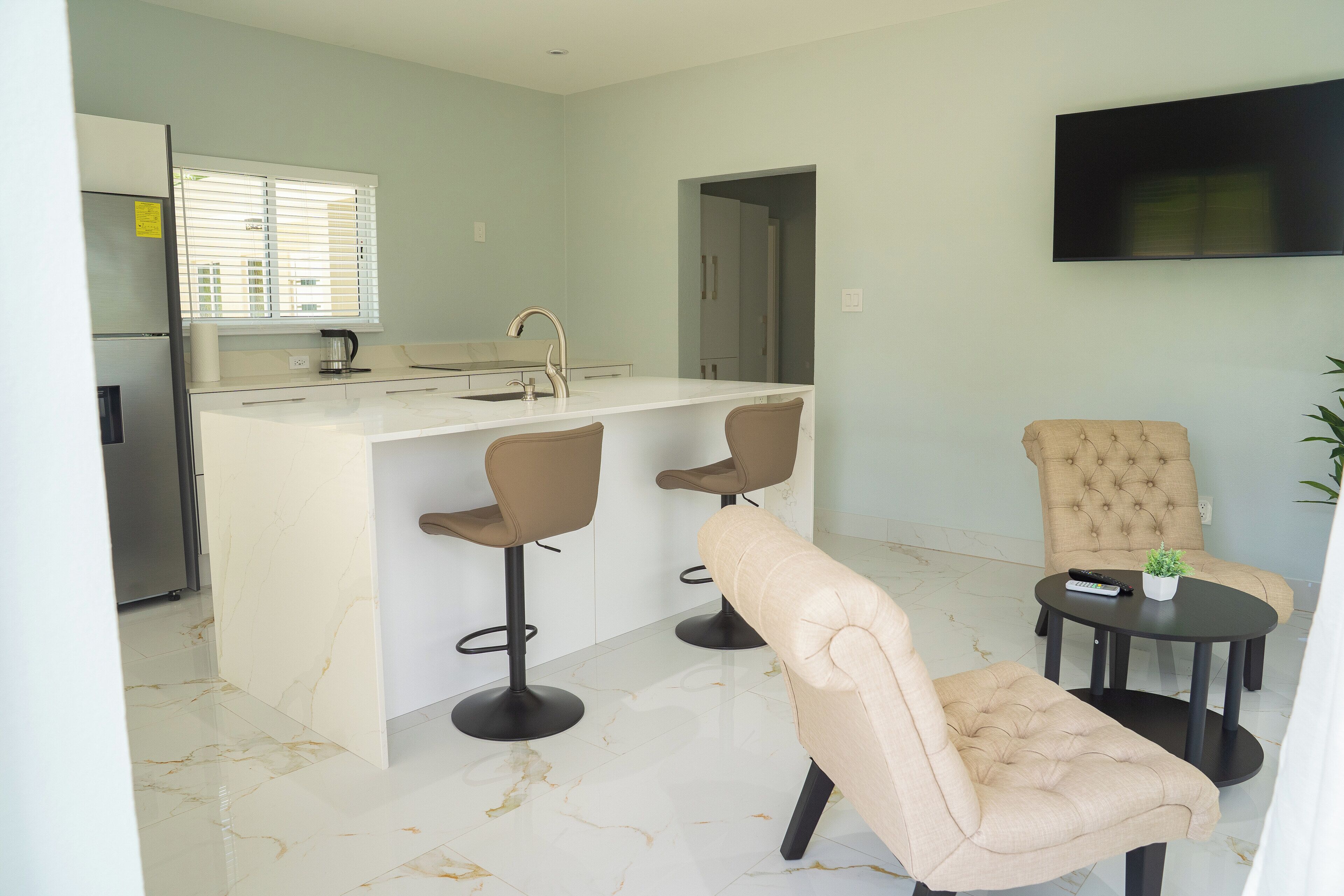 Una cocina moderna con una isla central, dos taburetes de bar, un sillón color crema y una pequeña mesa con una planta y un control remoto.