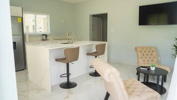 Una cocina moderna con una isla central, dos taburetes de bar, un sillón color crema y una pequeña mesa con una planta y un control remoto.