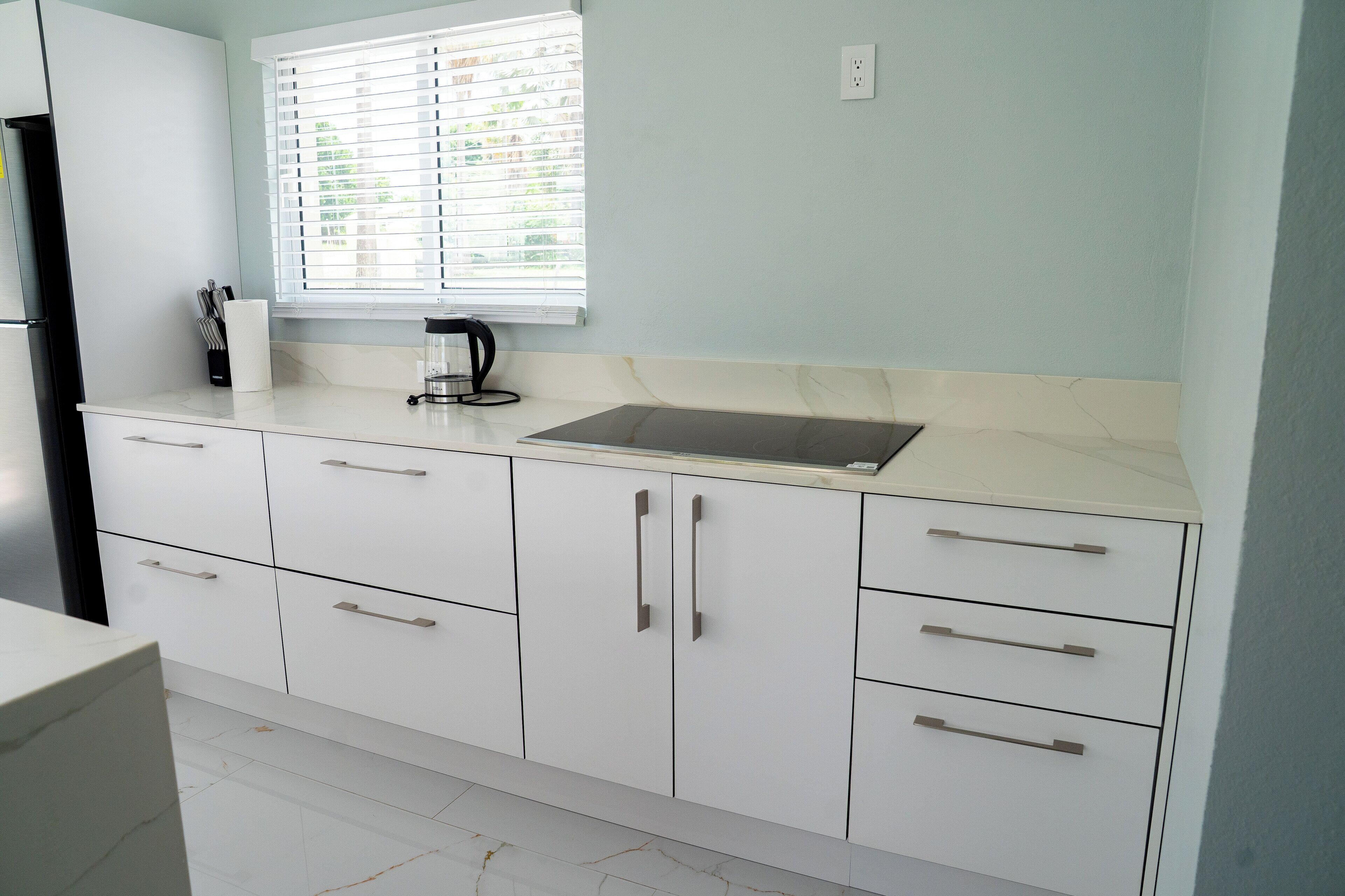 A kitchen with white cabinets, a window with blinds, and a countertop with a coffee maker.