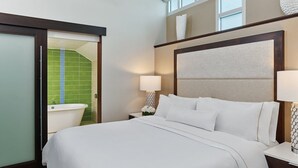 Habitación de hotel con cama, mesita de noche, lámpara, ventana y zona de ducha con azulejos verdes.