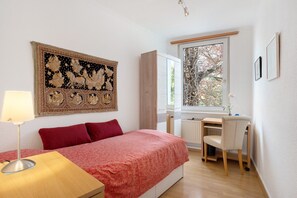 A bedroom with a bed, desk, chair, lamp, radiator, and a large tapestry.