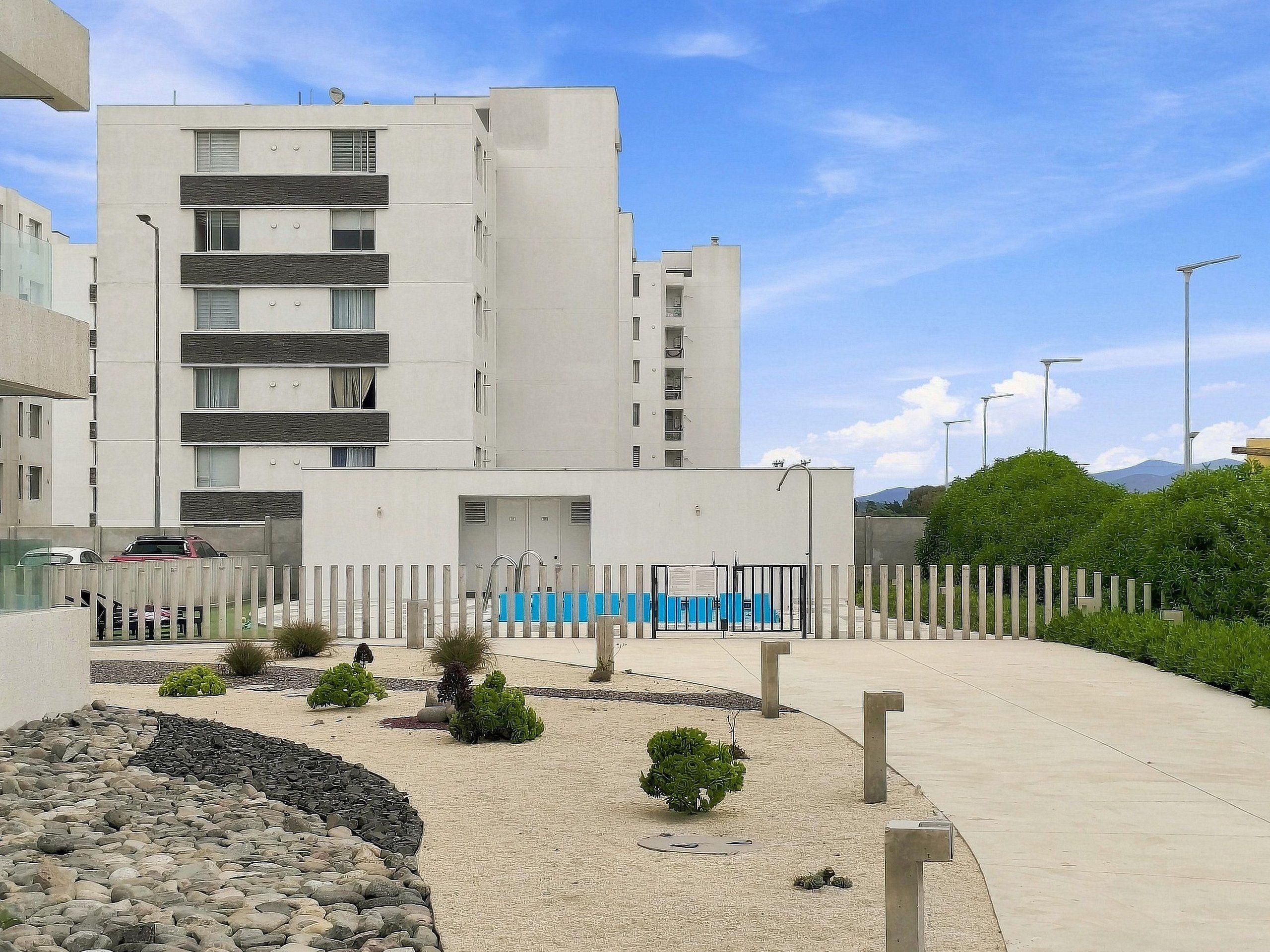 Un edificio moderno con varios pisos, una entrada con portón y un área ajardinada con rocas y arbustos.