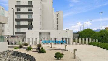 Un edificio moderno con varios pisos, una entrada con portón y un área ajardinada con rocas y arbustos.