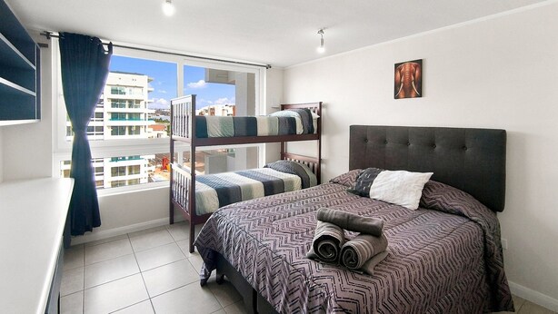 A bedroom with bunk beds, a large bed with patterned bedding, a window with curtains, and a city view. - Oceana Suites Puerto Serena (La Serena)