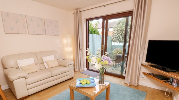 A living room with a sofa, a coffee table with a book and a vase of flowers, a TV on a stand, and a sliding glass door leading to a garden. - Poole Quay Cottage (Poole)