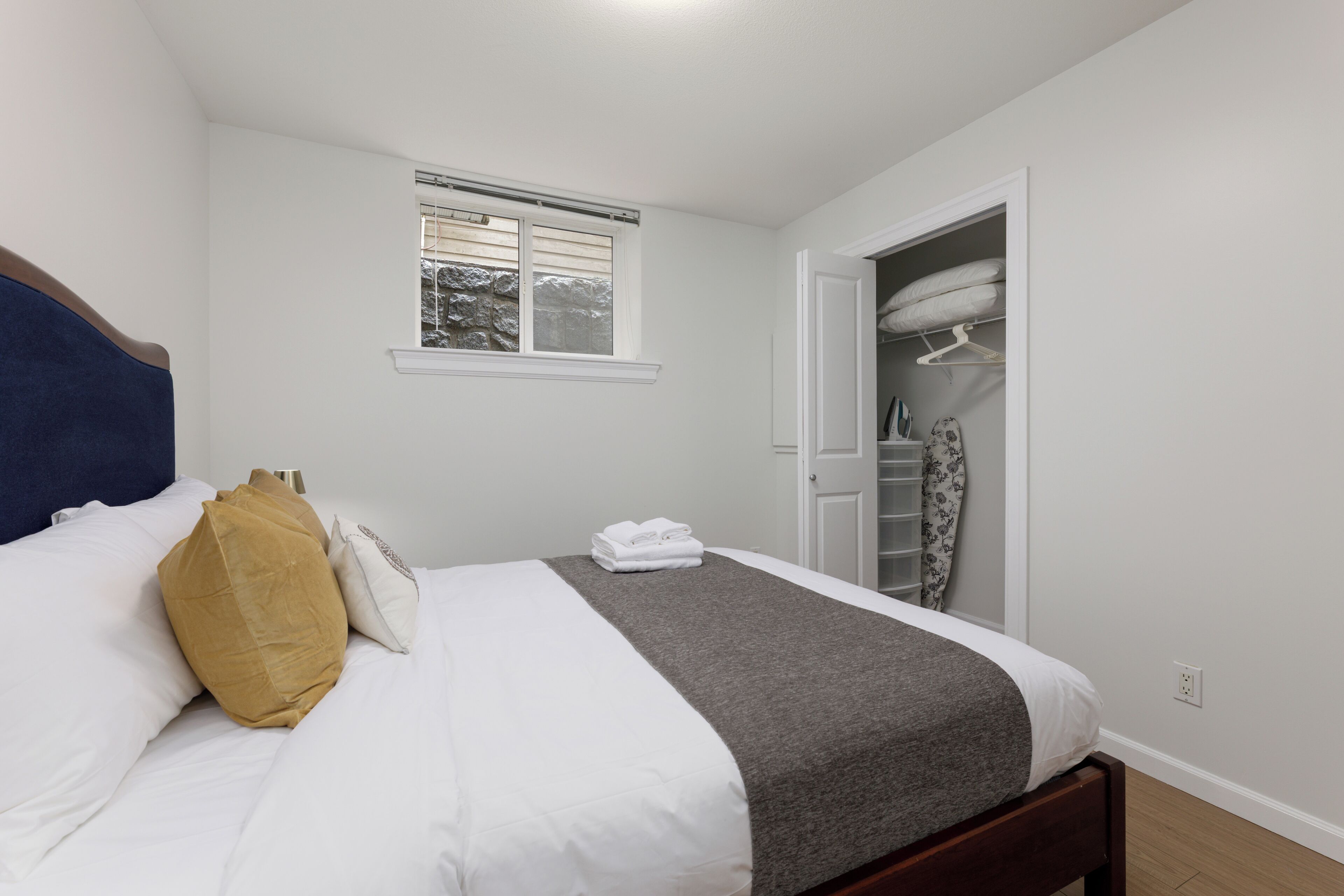 A neatly made bed with white and gray bedding, a wooden headboard, and a window with blinds.