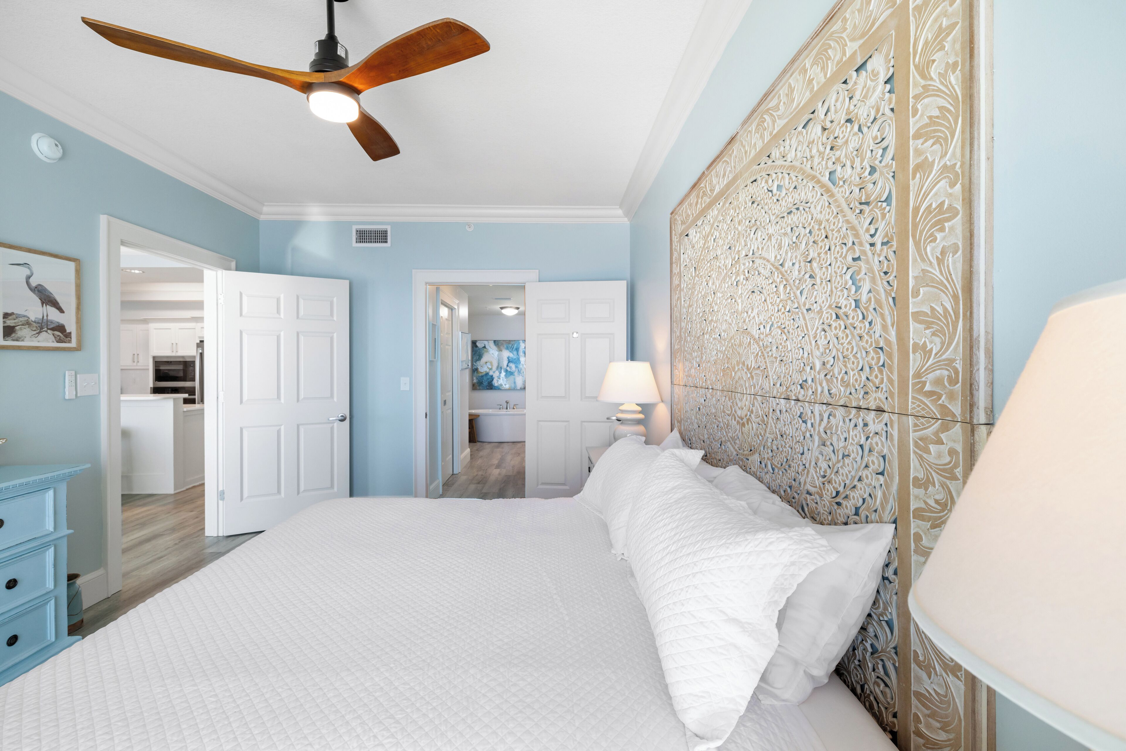 A bedroom with a large bed, a ceiling fan, a decorative headboard, and a view into the bathroom.