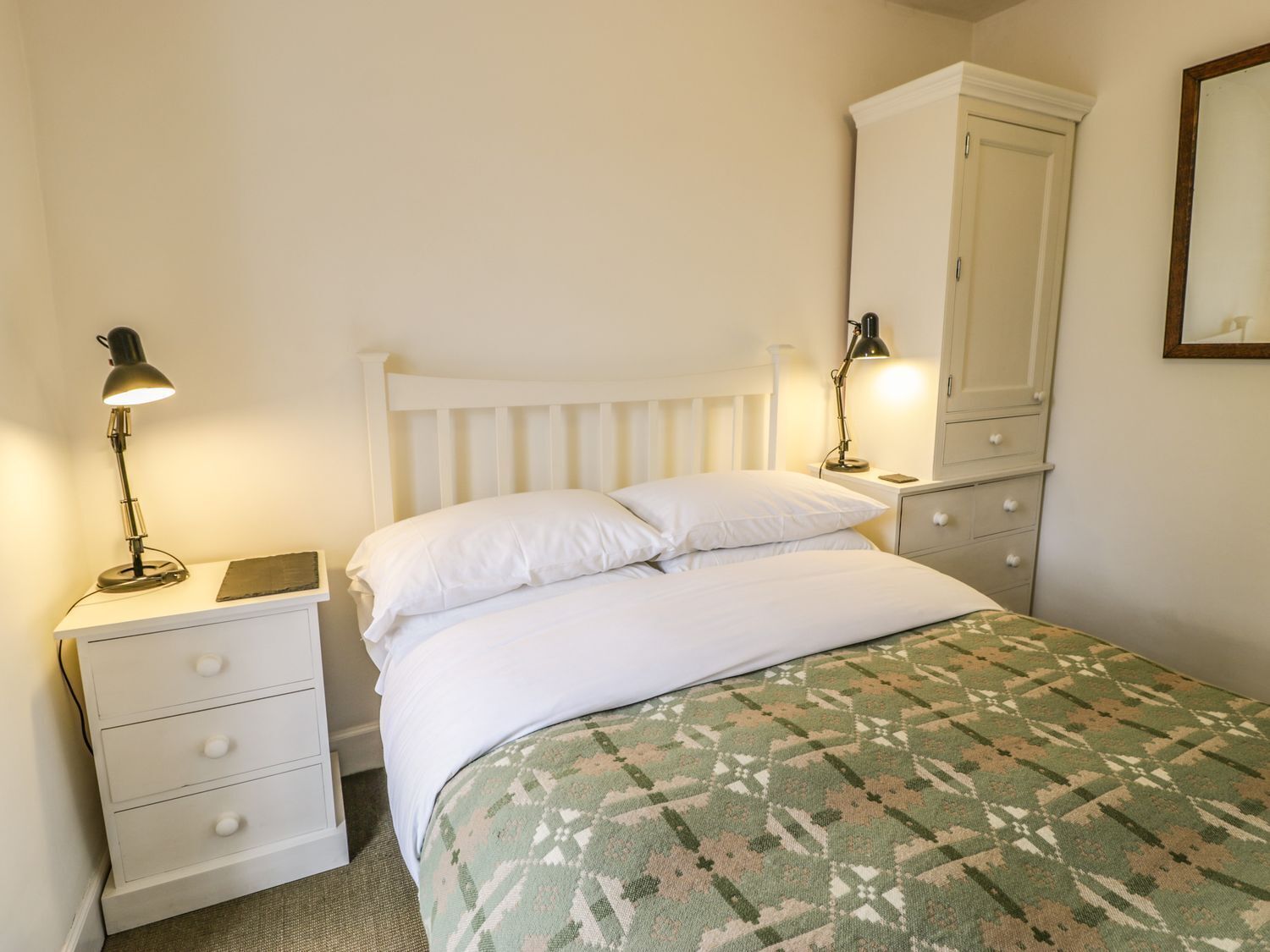 Une chambre avec un lit blanc, deux tables de chevet avec des lampes, une armoire blanche et un tapis à motifs.