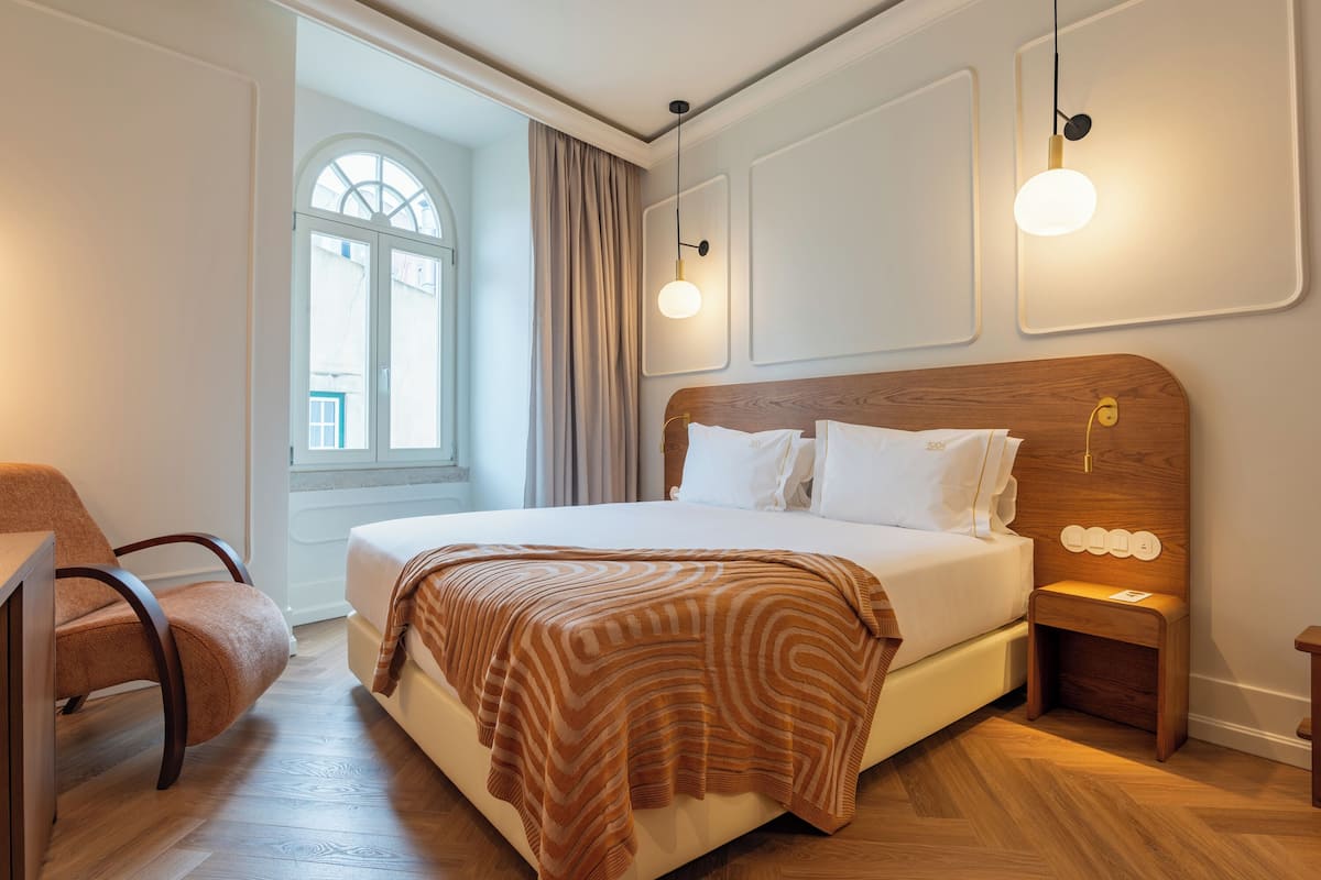 Ein Hotelzimmer mit einem großen Bett, einem Holfnachtkästchen, einem Sessel und einem Fenster mit Vorhängen.