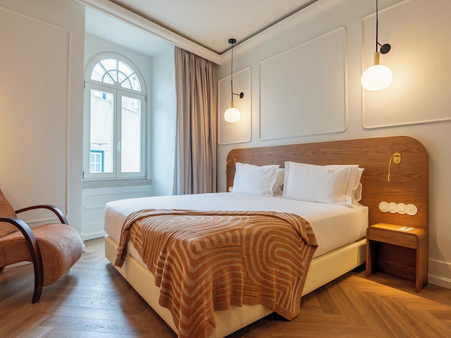 Hotellihuone, jossa on suuri sänky, puinen yöpöytä, tuoli ja ikkuna, jossa on verhot.