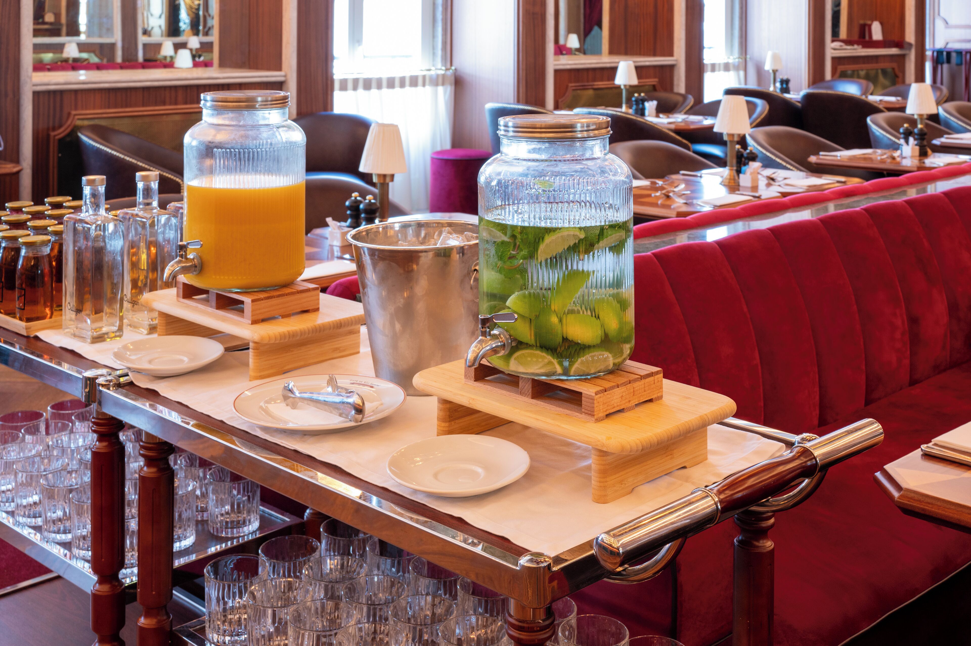 Un espace bar avec un long comptoir en bois, de nombreux verres, une grande carafe de jus d’orange et une grande carafe d’une boisson verte contenant des feuilles de menthe.