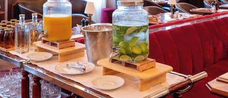 Un espace bar avec un long comptoir en bois, de nombreux verres, une grande carafe de jus d’orange et une grande carafe d’une boisson verte contenant des feuilles de menthe.