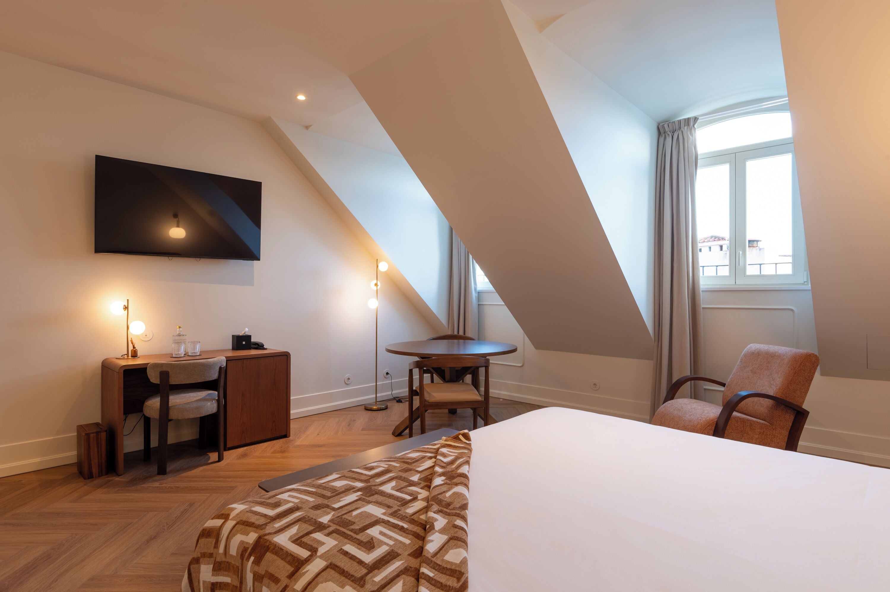 Une chambre d’hôtel moderne avec un grand lit, un bureau avec une chaise, une petite table, un téléviseur fixé au mur et une fenêtre avec des rideaux.