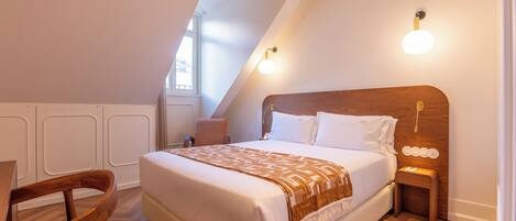 A bedroom with a large bed, a wooden headboard, a bedside table with a lamp, a chair, and a window with natural light.