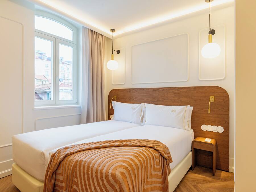 Een tweepersoonsbed met houten hoofdbord, twee nachtkastjes met lampen, een raam met gordijnen en een uitzicht op gebouwen.