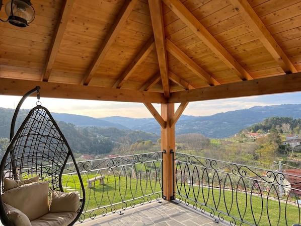 A wooden balcony with a hanging chair, a stone floor, and a metal railing.