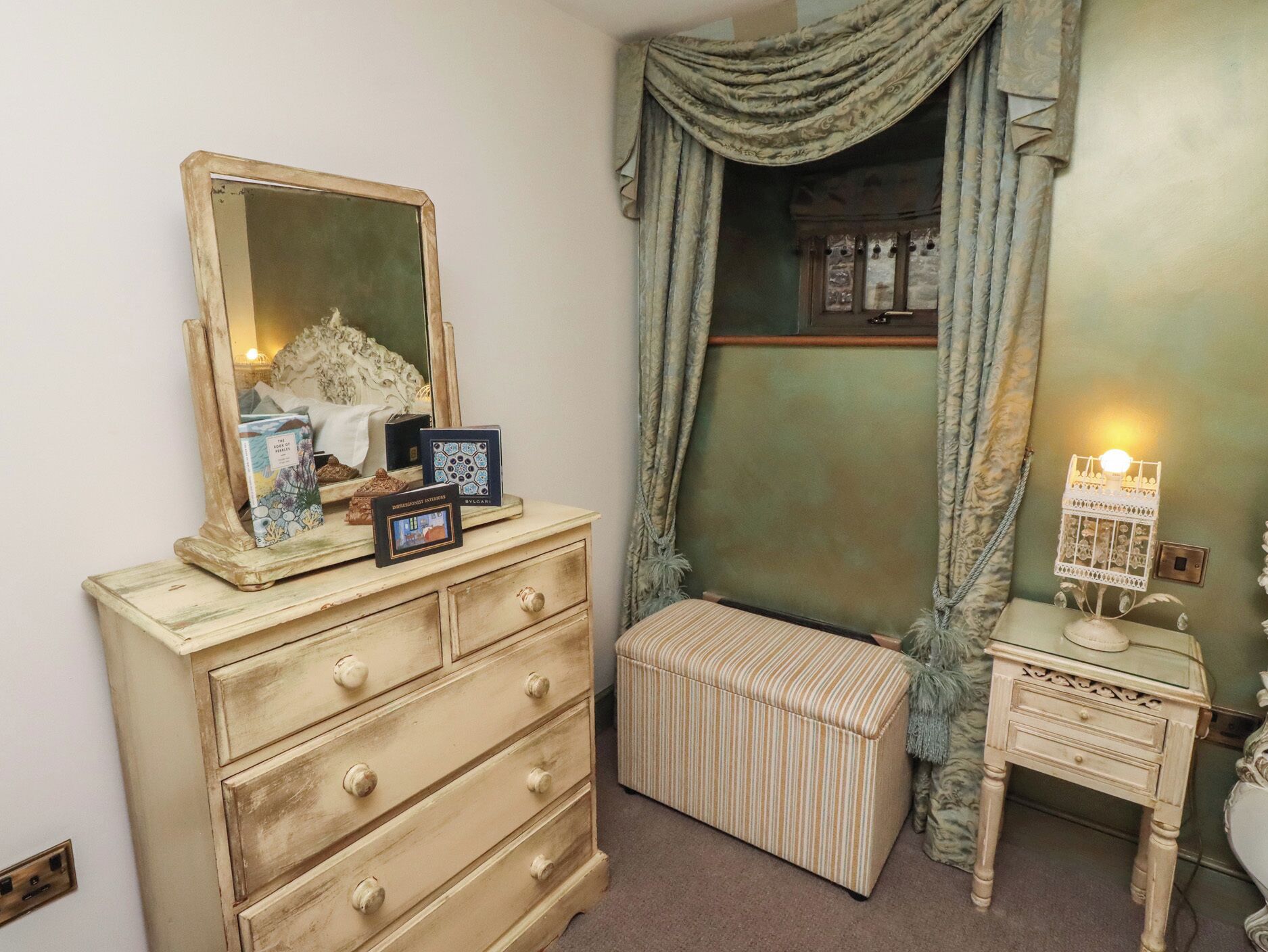 A bedroom with a dresser, a mirror, a bedside table, a lamp, and a window with curtains.