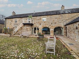 A stone building with a staircase leading to a balcony, a bench, and a wheelbarrow in the courtyard.