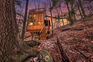 A wooden cabin with a staircase leading to a deck, surrounded by trees and fallen leaves. - The Hygge Treehouse Hocking Hills (Rockbridge)