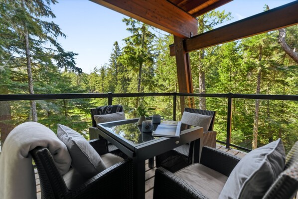 A balcony with wicker furniture, a glass table, and a view of a forest.