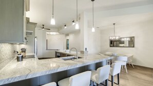 A modern kitchen with a marble countertop, bar stools, and pendant lighting. - Pelican Bay Retreat | Seasonal Resort Living in Pebble Creek (Naples)