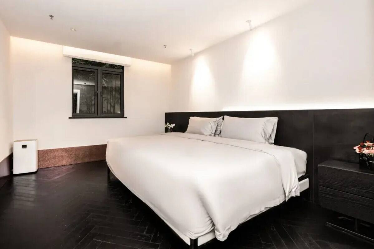 Un dormitorio moderno con una cama grande, una mesita de noche pequeña, una ventana y un suelo de madera oscura.