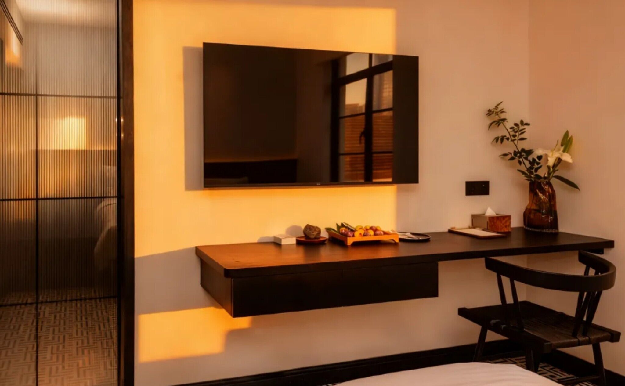 A hotel room with a wooden desk, a mirror, a vase with flowers, and a chair.